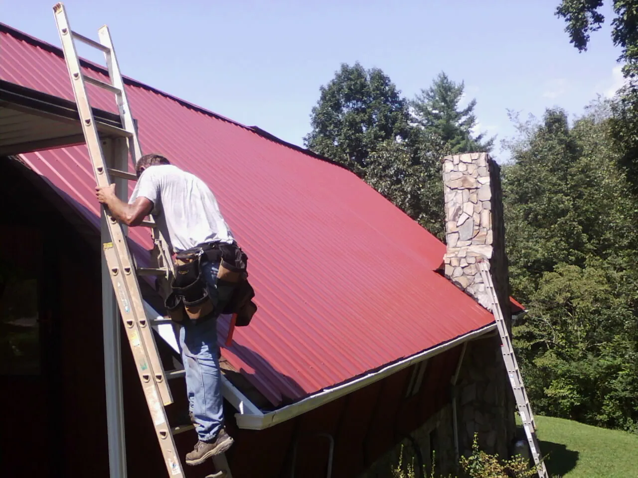 Roofer on a ladder beside a red metal roof and stone chimney — Soffit & Fascia Repair by Roofing Brothers in Moorestown